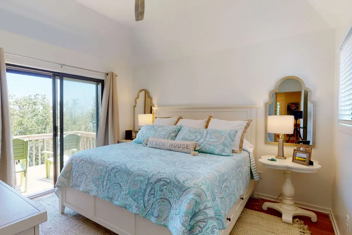 A serene bedroom with a bed dressed in blue paisley bedding, a decorative 'BEACH HOUSE' sign above the headboard, matching nightstands with lamps, and a prominent decorative mirror. The space exudes a relaxing, airy atmosphere and welcomes plenty of natural light.
