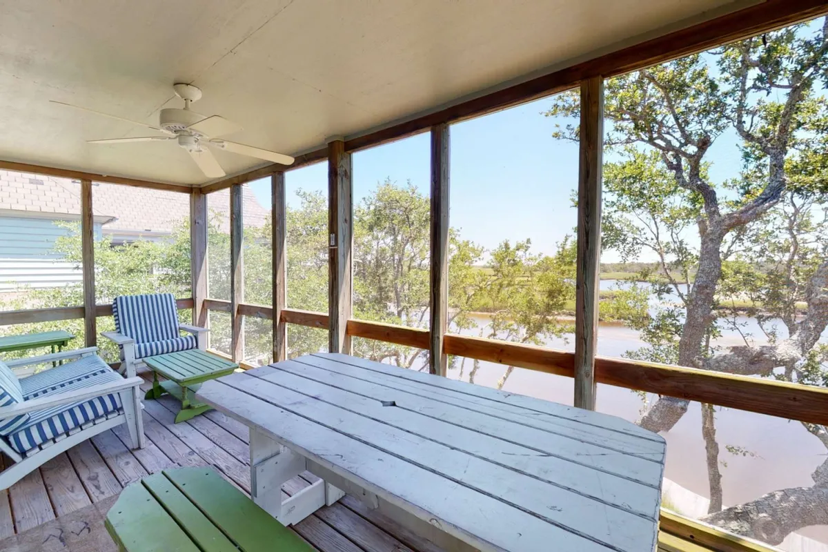 A wooden dining table with striped chairs is set in front of expansive windows that offer outdoor views. The scene is soaked in natural light and suggests a seamless transition from kitchen to dining and relaxation.