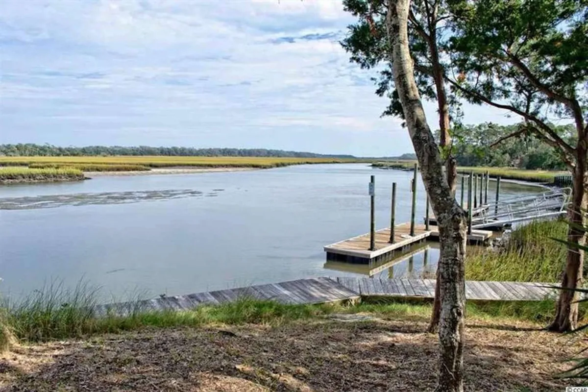A serene dock extends over a peaceful waterway, surrounded by lush trees and expansive marshland. The scene is quiet, inviting guests to fish, launch a kayak, or simply enjoy the natural beauty. The tranquil water under a soft sky captures the spirit of marshside adventure.