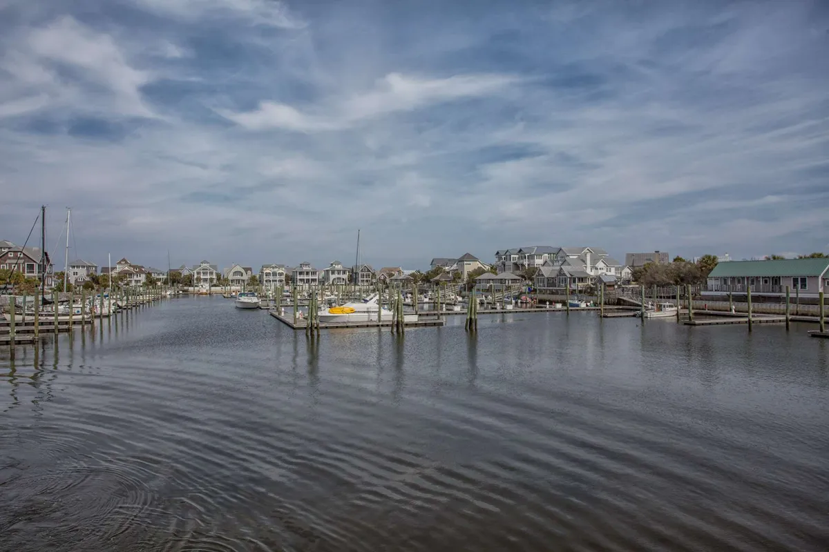 A marina scene with calm water, boats docked along the shore, and houses in the background. People can be seen boating, fishing, and enjoying the tranquil ambiance at the water’s edge.