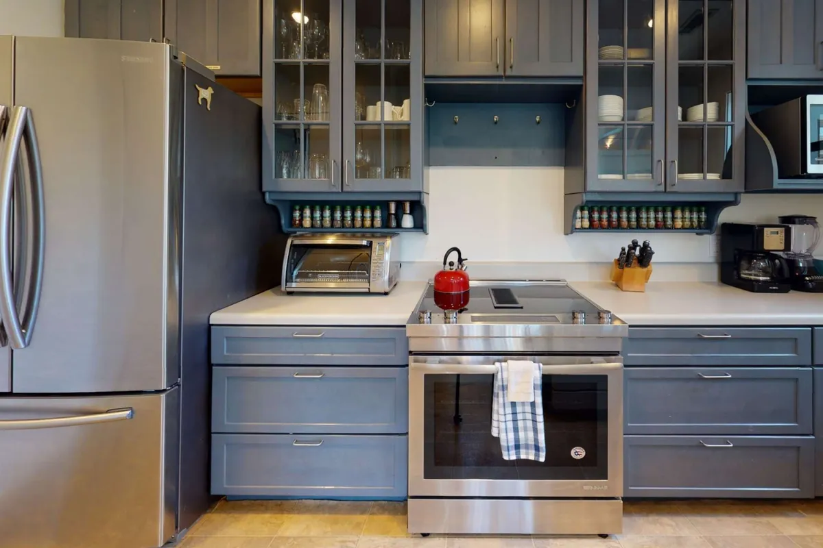 A bright, modern kitchen featuring stainless steel appliances, a red kettle adding a pop of color, and glass-front cabinets. The countertops are neatly arranged, and the space feels inviting for cooking, baking, and gathering.