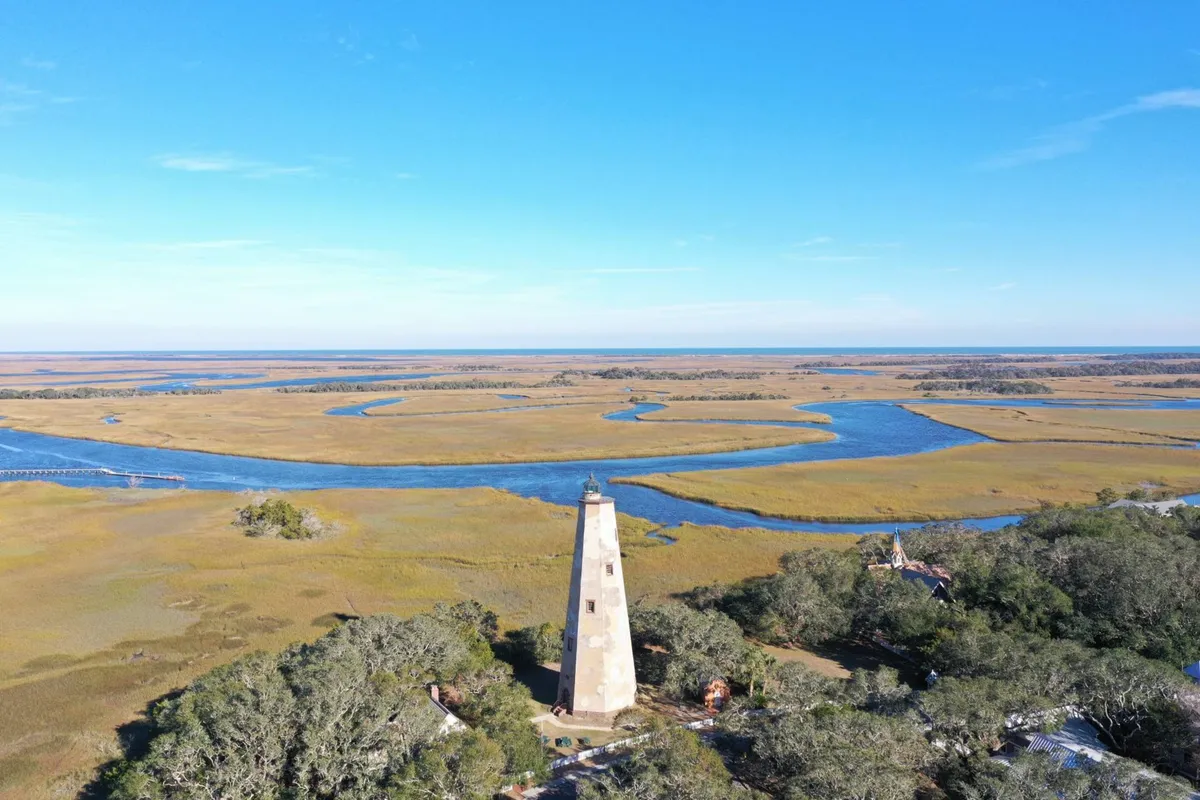 This image captures a sweeping view of the marshland, meandering waterways, distant horizon, and the iconic lighthouse. The tranquil landscape is dotted with native vegetation, providing a rich habitat for local birds and wildlife, making it an ideal visual representation of the marshfront setting described in the section.
