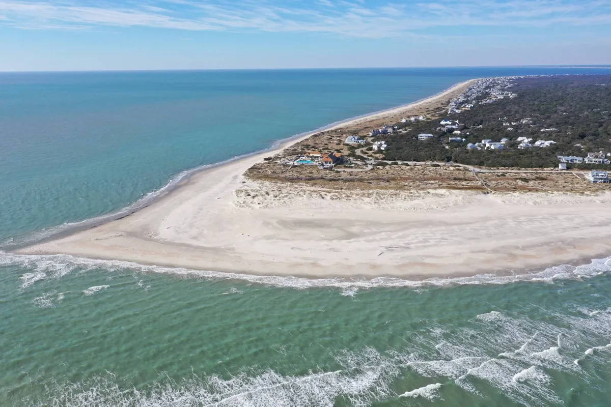 A wide sandy beach stretches along the coastline, with gentle waves lapping at the shore and distant houses dotting the horizon. The peaceful setting invites visitors to relax, beachcomb, swim, or simply take in the ocean views.