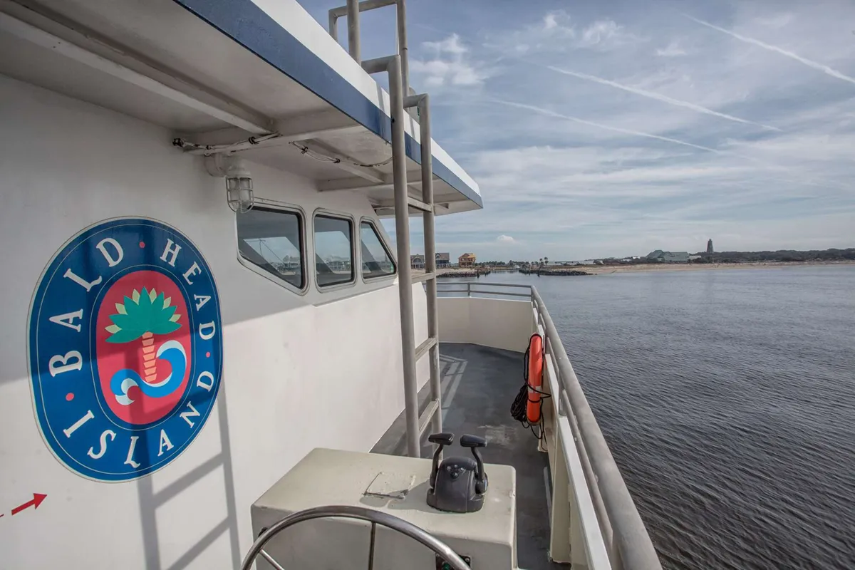 A boat deck adorned with the Bald Head Island logo, overlooking a serene river. The image captures the sense of transit and anticipation that comes with arriving on the island, with hints of adventure and seclusion. The perspective from the deck suggests the ferry-only access that defines the experience.