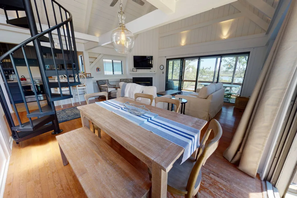 This image presents an inviting open-concept interior with a visible spiral staircase, spacious dining table, ample comfortable seating, and large windows that draw in natural light. The windows offer glimpses of the marsh views beyond, while the arrangement of furniture fosters interaction and communal experiences.