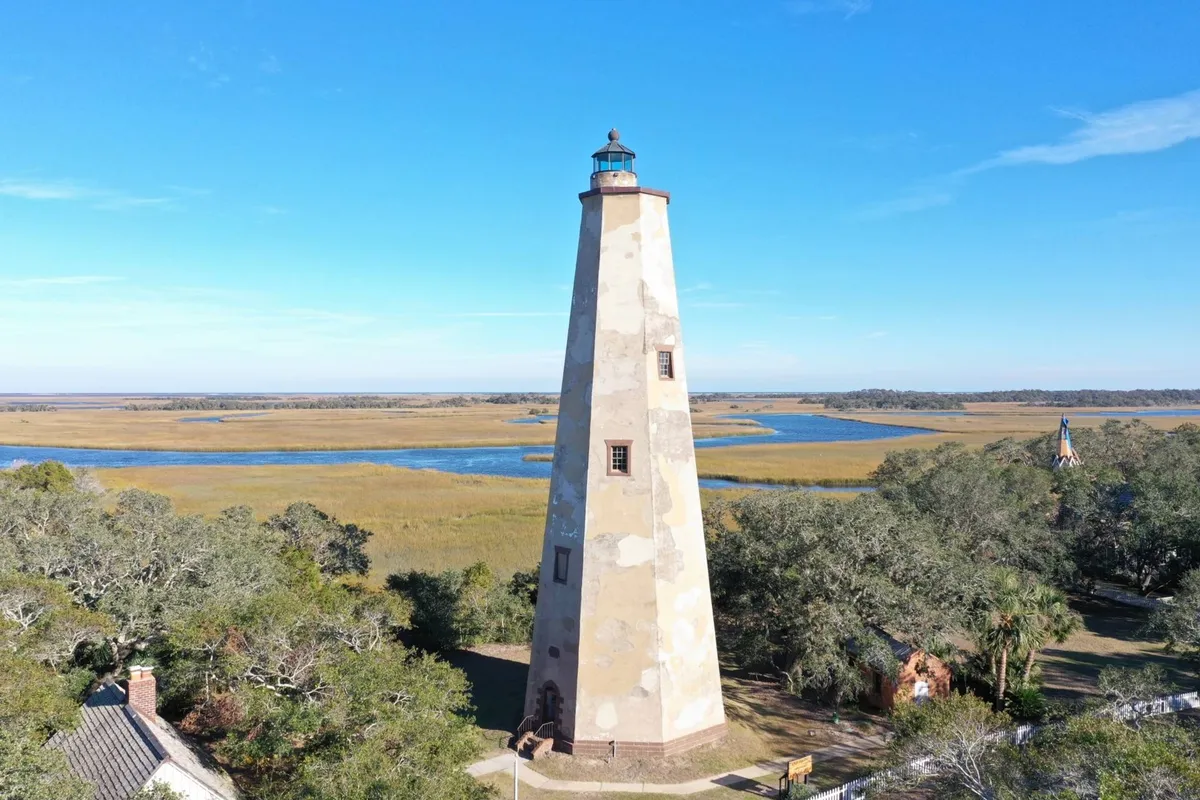 A picturesque lighthouse sits beside lush marshland and a tranquil waterway under a bright blue sky. The scene evokes the island’s natural and historic charm, inviting visitors to explore its iconic landmarks and scenic landscapes.