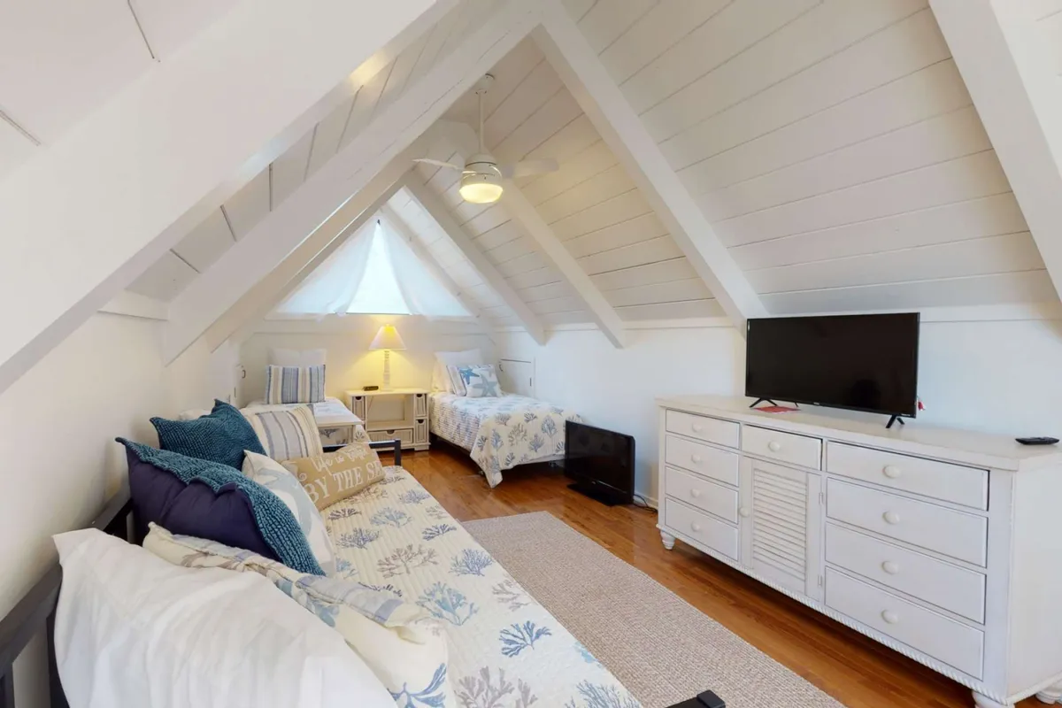 A charming loft space with a sloped ceiling, patterned bedding on twin beds, a trundle bed, and soft lighting from stylish lamps. A TV unit and wooden floors add functionality, making this a playful hideout for kids or a quiet retreat for reading and relaxation.