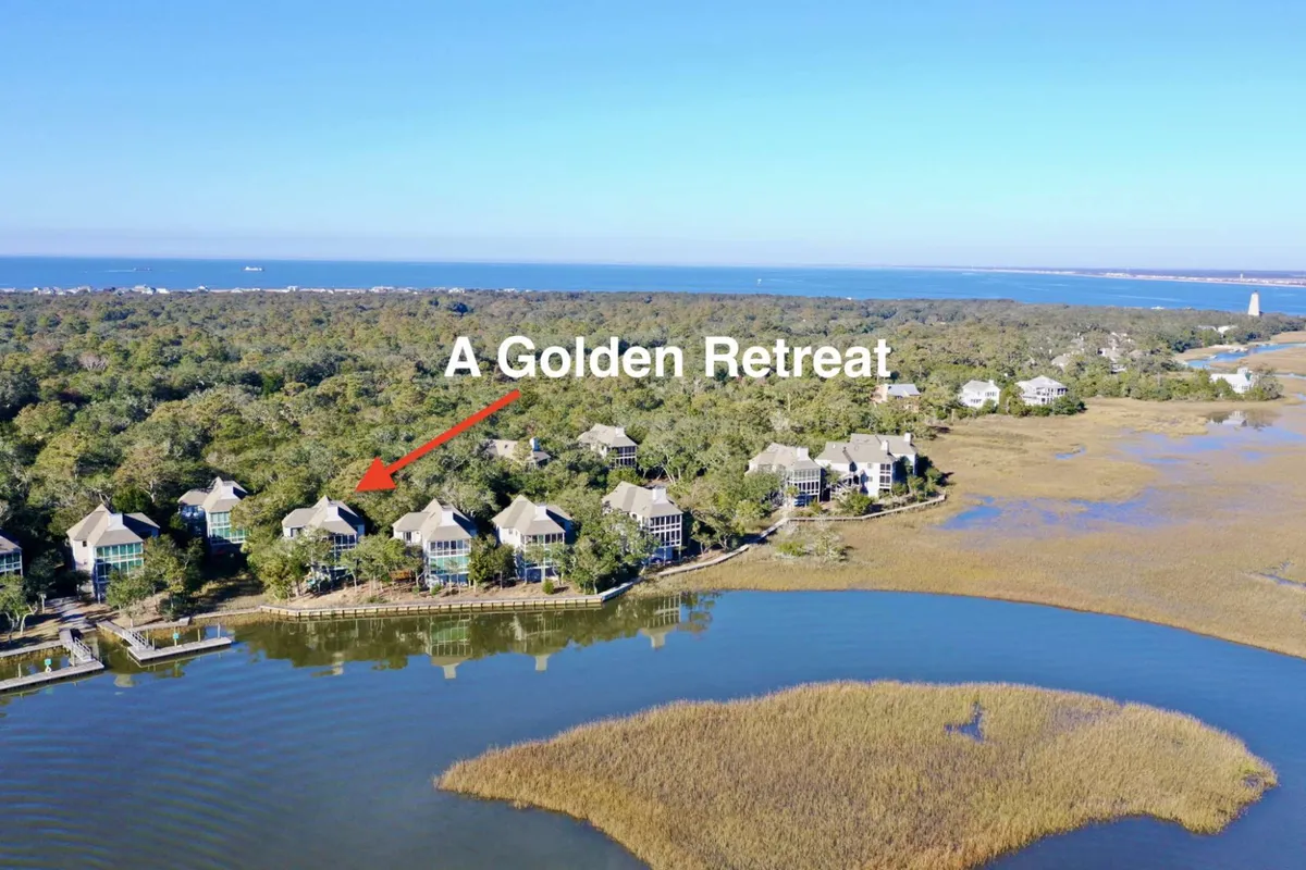 A peaceful marshfront view featuring open water, lush greenery, and a golden retreat house in the distance. The calm setting is ideal for kayaking and observing local wildlife, with soft light suggesting either sunrise or sunset.