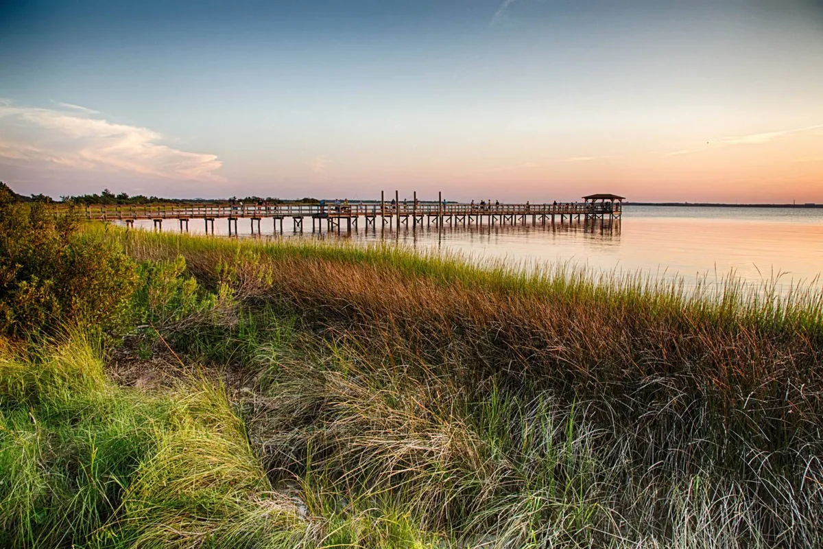 A serene wooden pier extends into calm water lined with tall marsh grasses, under a glowing sunset sky. The setting feels inviting for fishing, strolling, and watching the evening colors reflect on the water.