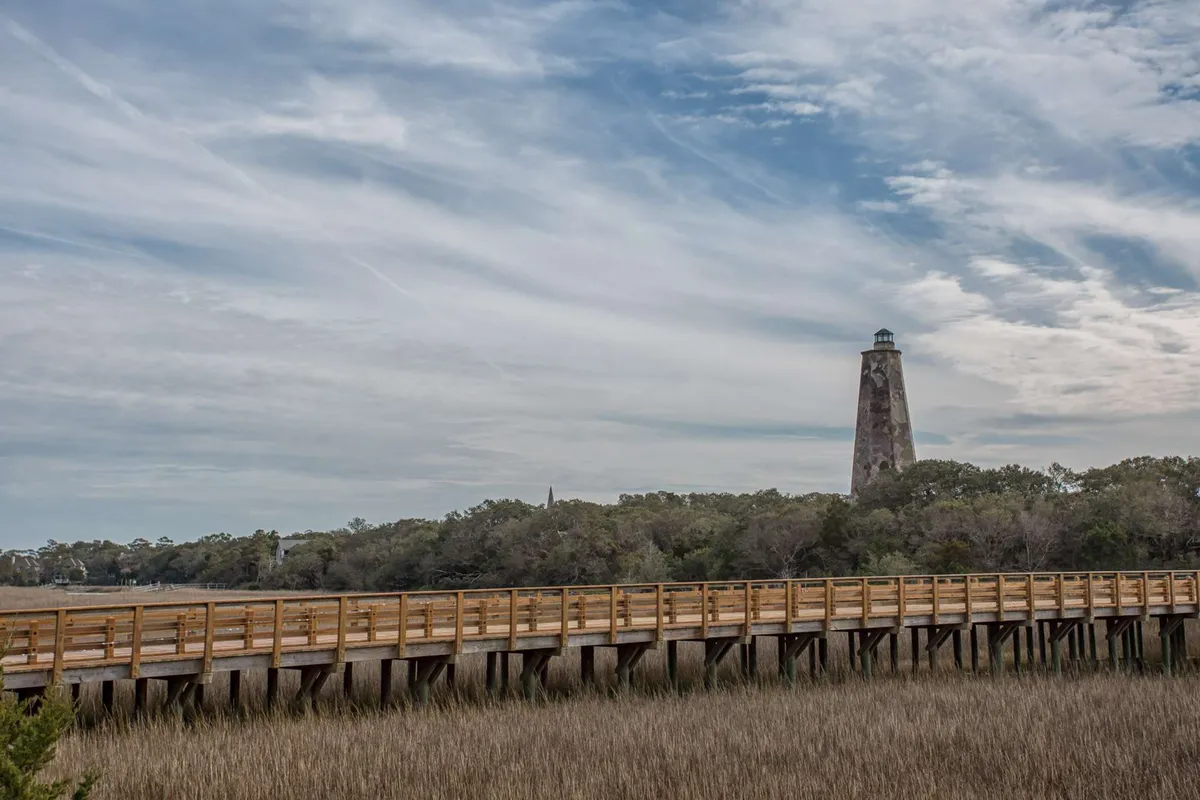 A wooden walkway leads out into the marshland, with the lighthouse visible in the background. Grassy wetlands surround the path, inviting exploration and hinting at the wildlife and birds that frequent this area. The calm mood and open vistas encourage viewers to imagine themselves venturing out for birdwatching or photography.