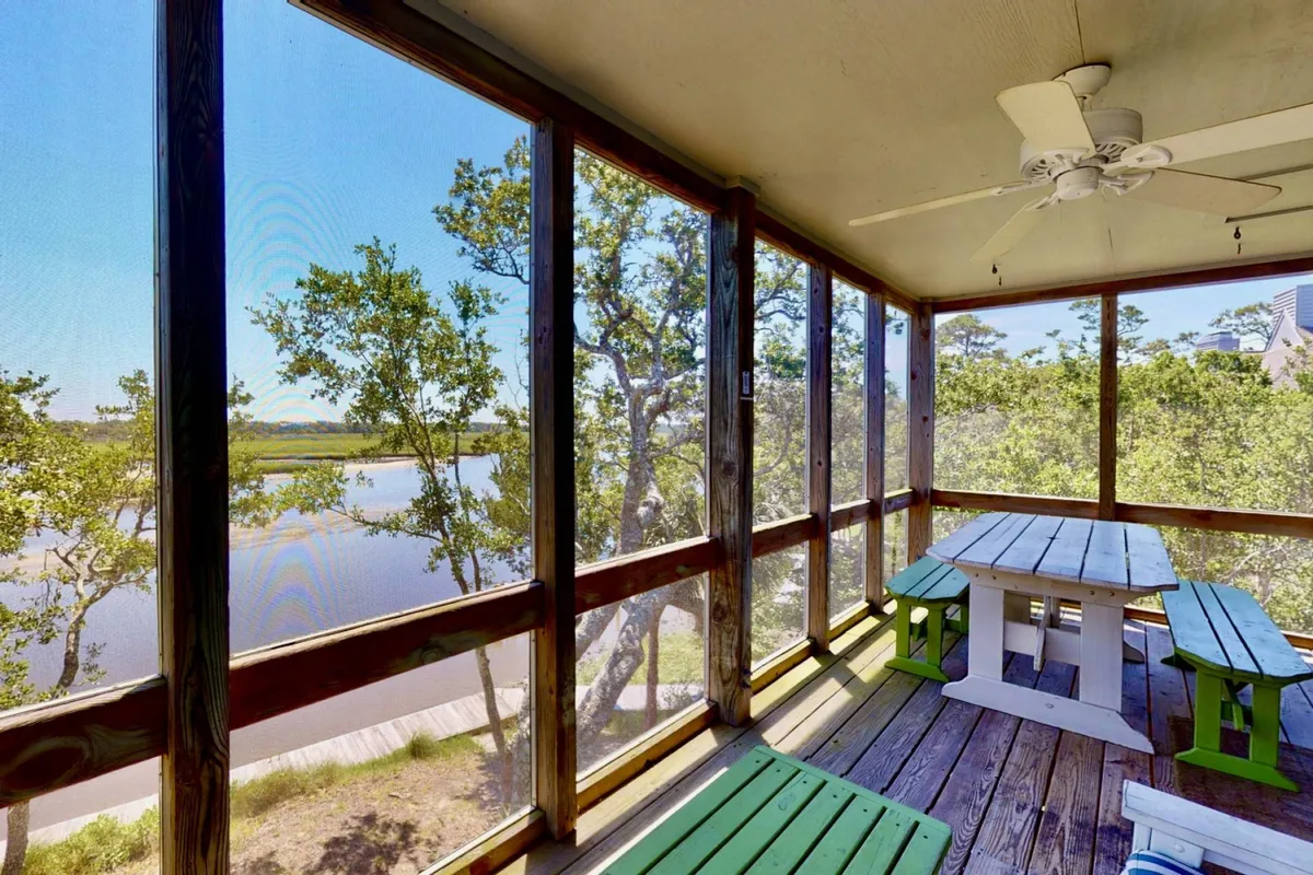 The image showcases a bright, screened porch with a dining table surrounded by cheerful green chairs overlooking the tranquil waters of Bald Head Creek and Salt Marsh Sanctuary. The inviting setup hints at leisurely meals, peaceful mornings, and opportunities to watch wildlife right from the comfort of the condo.