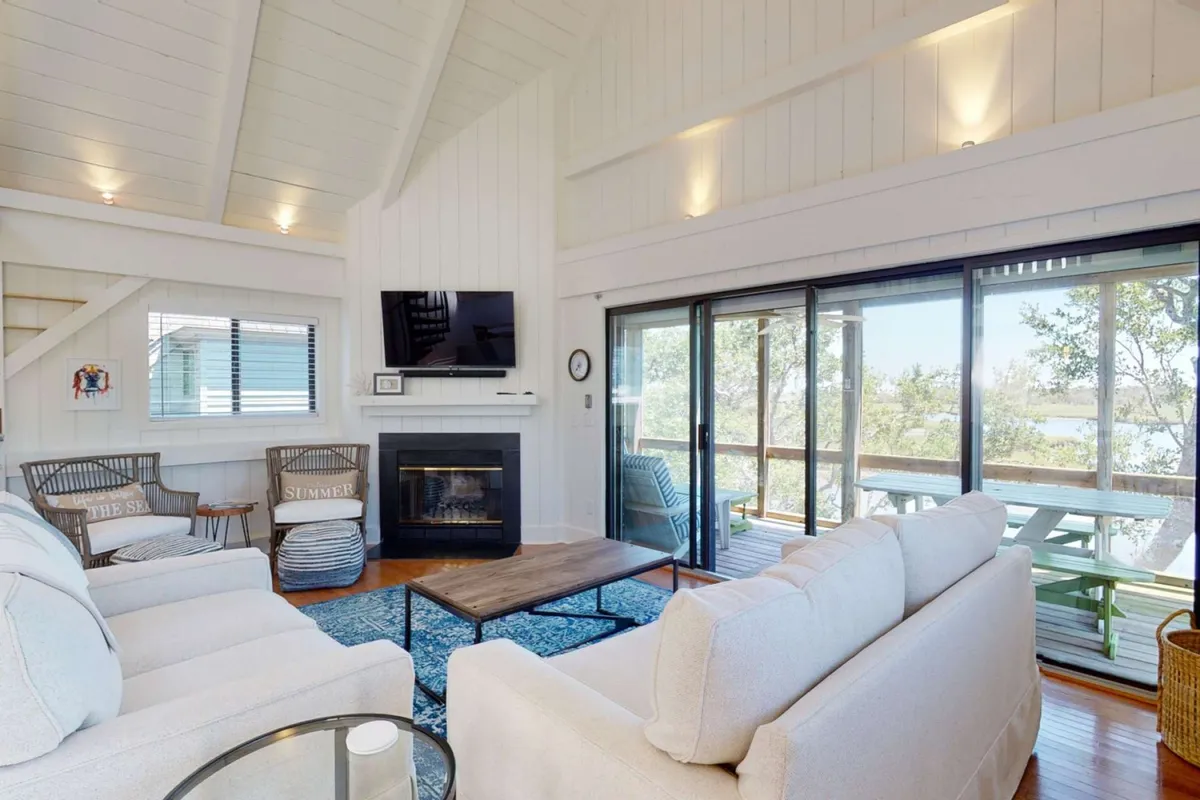 The scene features a comfortable sofa set grouped around a fireplace, with large windows letting in daylight and hints of the natural surroundings outside. Decorative touches and a plush rug create a welcoming environment ideal for relaxing, movie nights, or enjoying the marsh scenery.