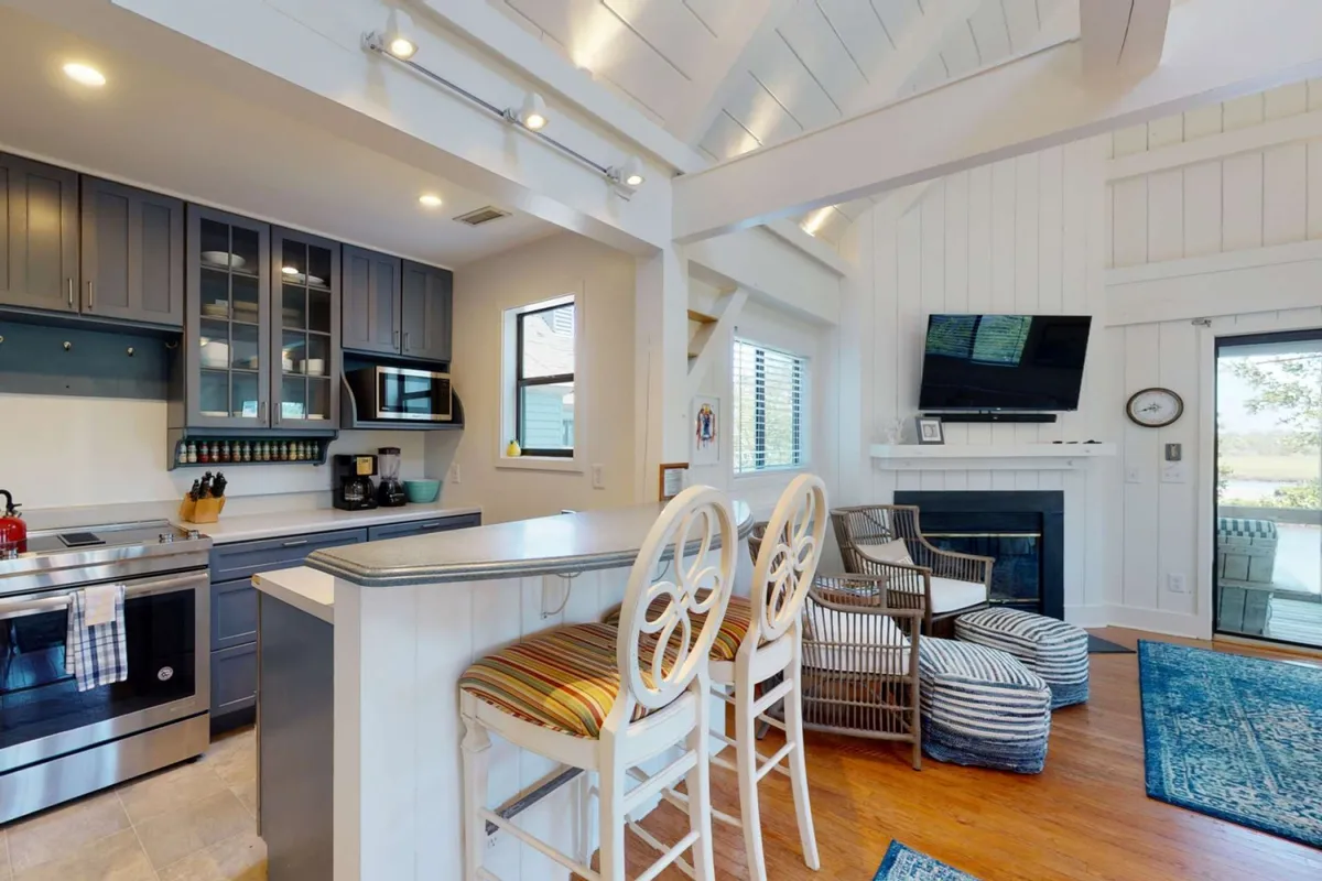 An open kitchen layout with a central island and bar stools, adjacent seating area, fireplace, and large windows that let in natural light. The space encourages socializing, cooking, and enjoying views of the outdoors.