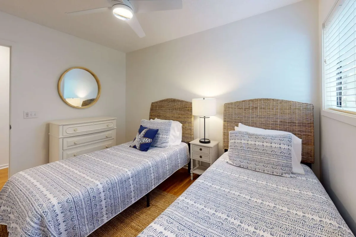 A cozy bedroom featuring two twin beds with decorative pillows, a nightstand with a lamp positioned between them, and a dresser with a mirror above. The space feels bright, calm, and kid-friendly, ideal for siblings, teens, or grandparents.